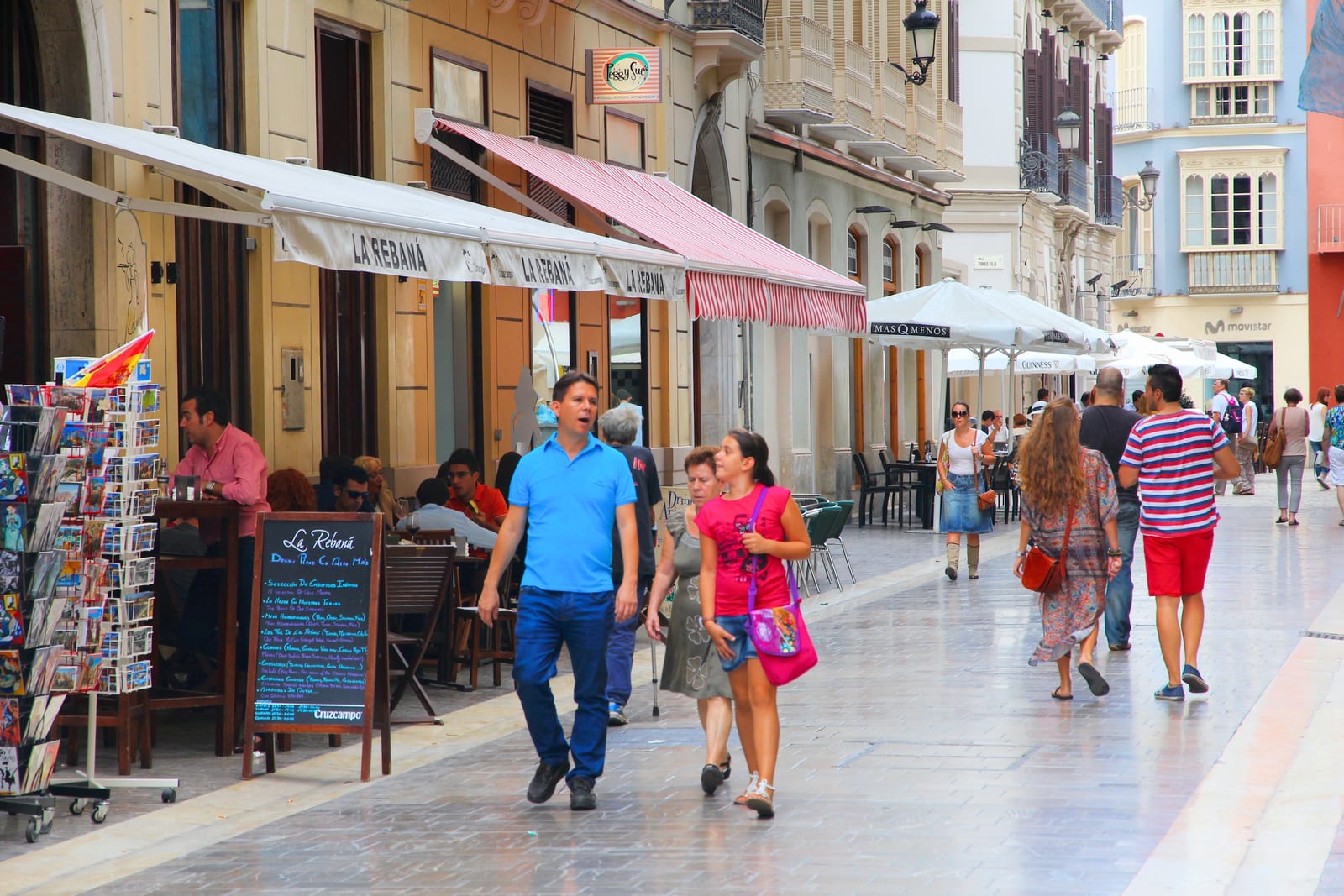 Pedestrians on a street in central Málaga, with restaurant awnings and a postcard stand