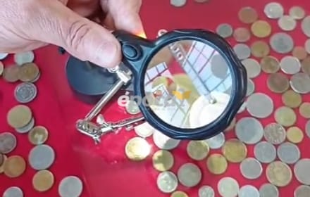 A hand with a loupe over coins of different periods on a red surface