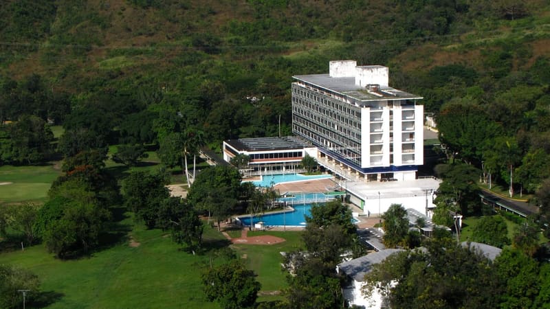 Vista aérea del Hotel de Golf Maracay, edificio blanco de varios pisos con piscinas y campo de golf al pie de las montañas de Aragua