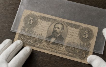 Vintage five-denomination banknote with an oval portrait and engraved borders, held with cotton gloves while being slid into a clear protective sleeve on a wooden table