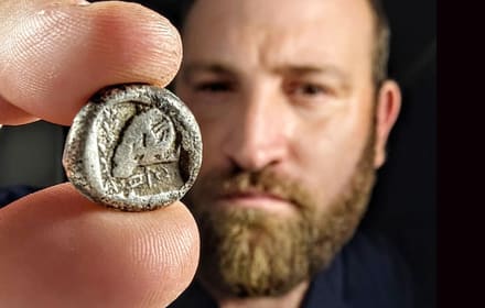 Ancient Cypriot silver coin (Salamis) held between fingers, with a ram relief; a researcher in the background