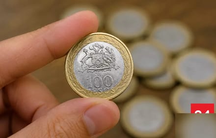 A hand holds a Chilean bimetallic 100-peso coin with the national arms over other coins